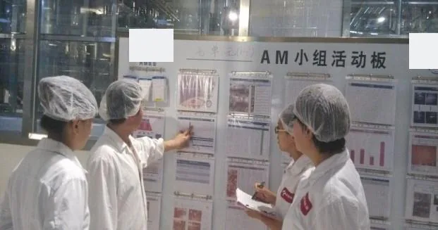 Operators in white coats and hairnets reviewing an autonomous maintenance team activity board on the production floor.